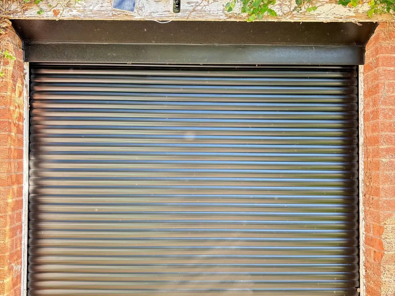 Dark grey aluminium roller shutter fitted in a brick opening below an ivy-covered lintel